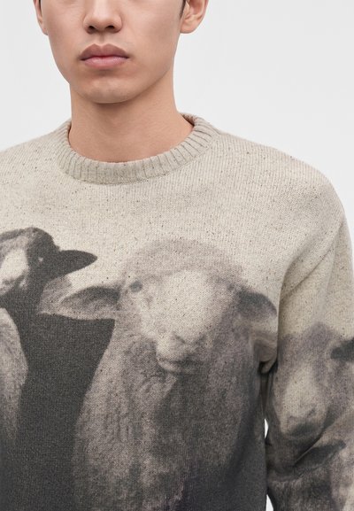 Homme portant un pull beige avec des têtes de moutons noires et grises imprimées sur le devant sur un fond clair uni.