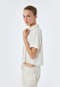 Korte mouwen, overhemd met kraag in lichtbeige stof. Vrije pasvorm met een iets verkorte zoom, gecombineerd met bijpassende beige sweatpants met trekkoord.