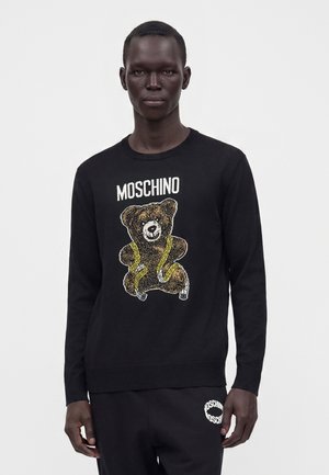 Mężczyzna noszący czarny sweter Moschino z nadrukiem misia i miarki, w połączeniu z czarnymi spodniami Moschino, stoi na białym tle.