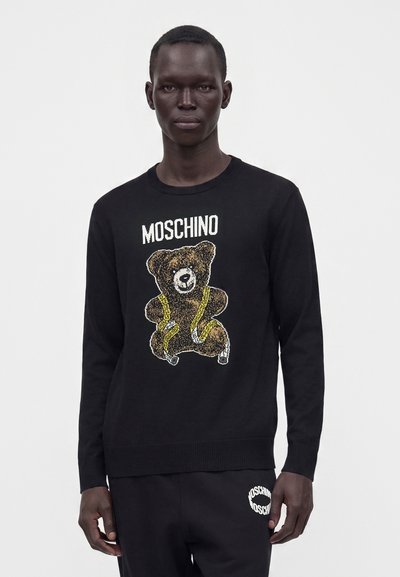 Homme portant un pull noir Moschino avec un motif d'ours en peluche et de ruban à mesurer, assorti à un pantalon noir Moschino, debout sur fond blanc.
