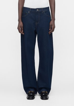 Tamnoplave traperice širokih nogavica koje nosi osoba tamne puti, uparene s crnim mokasinama i bijelim topom vidljivim u struku.