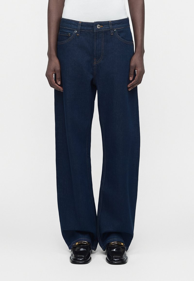 Σκούρο μπλε τζιν παντελόνι με φαρδιά μπατζάκια φορεμένο από άτομο με σκουρόχρωμο δέρμα, σε συνδυασμό με μαύρα loafers και λευκό τοπ ορατό στη μέση.