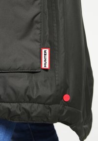 Černá nepromokavá bunda s náprsní kapsou s červenou nášivkou "HUNTER" a detailem se červeným patentním knoflíkem na spodní straně. Hladká textura.
