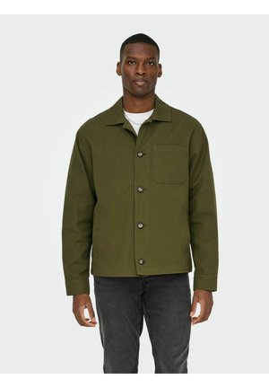 Man draagt een olijfgroene jas met knopen over een wit overhemd en donkergrijze spijkerbroek, staand tegen een effen lichte achtergrond.