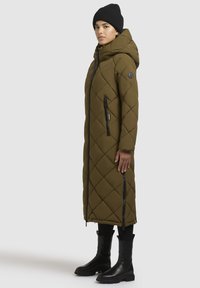 Lang olivengrøn pufferjakke med diamantquiltet mønster, lynlås foran og sidelommer med lynlås. Sammen med sorte ankelstøvler og en strikket hue.