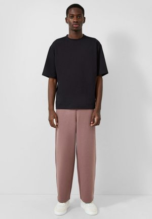 Młody mężczyzna stojący przodem, ubrany w czarną, oversize'ową koszulkę, luźne różowe spodnie, białe trampki i pojedyncze kolczyki.