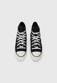 Tenisi negri cu bot din materiale albe și șireturi albe, văzuți dintr-o parte de sus pe un fundal deschis.