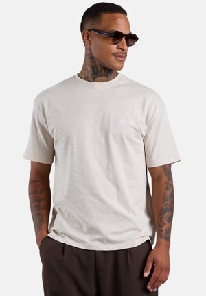 Mann mit Hals- und Arm-Tattoos, brauner Sonnenbrille, hellbeigem T-Shirt und dunkelbrauner Hose, schaut leicht nach rechts und hat die Hände in den Taschen.