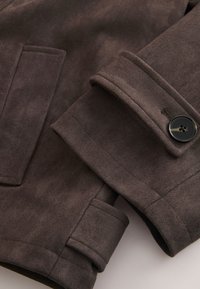 Nahaufnahme eines Ärmels einer braunen Wildlederjacke mit schwarzem, geknöpftem Manschettenverschluss und einer Seitentasche mit detaillierter Naht.