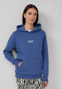Blauwe katoenen hoodie met een voorzak en witte geborduurde tekst "JOUW LICHAMEN IS JOUW GROOTSTE KAPITAAL." Draag met zwart-witte zebraprint broeken.
