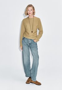 Vrouw met kort blond haar, gekleed in een beige trui, blauwe wijde jeans, bruine loafers en een lange zwarte ketting, staand tegen een witte achtergrond.