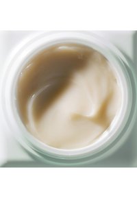 Hydratant crème beige dans un conteneur en verre rond, texture lisse visible, avec un éclat brillant. Pas de motifs distincts ni de matériel présent.