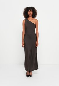 Robe maxi marron foncé à une épaule en tissu lisse et soyeux, ajustée au niveau du buste avec une légère évasement à l'ourlet. Chaussures à talons noires.