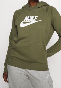 Olivgrön huvtröja med en stor vit Nike-logga, utrustad med en känguruficka, dragsko i huvan och ribbade muddar och fåll.