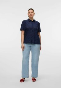 Camicia corta blu navy con colletto a bottoni, caratterizzata da un motivo floreale testurizzato e dettagli ritagliati. Indossata con jeans a gamba larga blu chiaro.