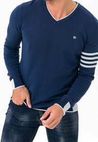 Marineblauer V-Ausschnittpullover aus geripptem Stoff mit weißen Streifenakzenten an den Ärmeln und kontrastierenden Bündchen. Schmale Passform.
