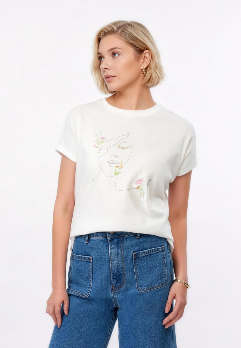 Ξανθιά γυναίκα που φοράει λευκό t-shirt με αφηρημένο σχέδιο γραμμής προσώπου με λουλούδι, μπλε τζιν με μεγάλες μπροστινές τσέπες, χρυσούς κρίκους στα αυτιά και βραχιόλι.