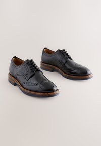 Black leather dress shoes featuring brogue detailing, lace-up closure, and a contrasting brown sole with a slight heel.