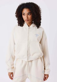 Cremfarbener Kapuzen-Zip-Hoodie mit blauen Akzenten, gerippten Bündchen und zwei Fronttaschen. Hergestellt aus weichem, strukturiertem Stoff.