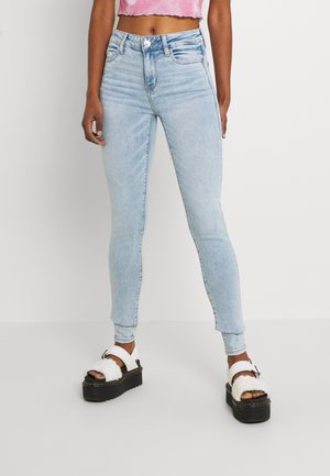 Light blue skinny jeans with a high waist and slight fading, paired with chunky white sandals featuring black soles and decorative straps.