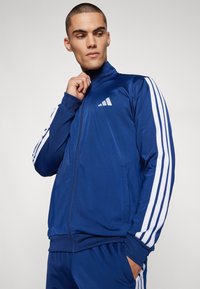 Modra zip-up trening jakna iz gladkega materiala, s belimi tridelnimi poudarki na rokavih in logotipom na prsih, visoko ovratnico in stranskimi žepi.