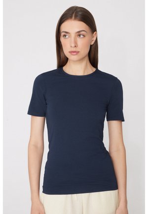 Donna con capelli castani lisci che indossa una maglietta aderente blu navy a maniche corte e pantaloni chiari, in piedi davanti a uno sfondo semplice.