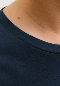 Gros plan sur le cou et la partie supérieure de la poitrine d'une personne portant un t-shirt en jersey côtelé bleu foncé avec un petit grain de beauté sur la peau visible.