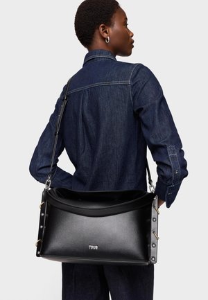 Kobieta w ciemnej dżinsowej koszuli niosąca dużą czarną skórzaną torebkę na ramię TOUS z metalowymi ćwiekami po bokach na jednolitym tle.