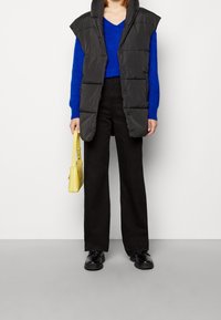 Person som bär en svart vadderad väst, blå V-hals tröja, svarta byxor, svarta skor och bär en liten gul handväska, stående på grått golv.