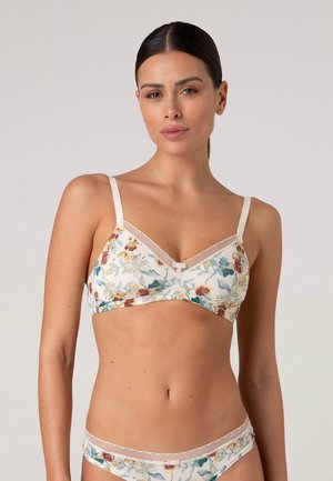 Donna con capelli scuri che indossa un reggiseno e biancheria intima floreali coordinati con spalline bianche e rifiniture in pizzo, in piedi davanti a uno sfondo semplice.