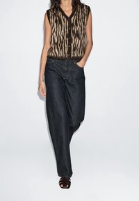Femme portant un gilet sans manches marron et beige à motifs avec des boutons, un jean large foncé et des sandales noires à bout ouvert, debout avec une main dans la poche.
