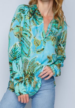 Kvinde i en bluse med teal og brune bladmønster med V-hals, iført lyseblå jeans, der holder blusens kant med den ene hånd.