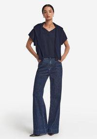 Marinblå topp med vida ärmar, lager på lager över en mörkblå skjorta, kombinerat med vida mörk denimjeans och svarta spetsiga skor.