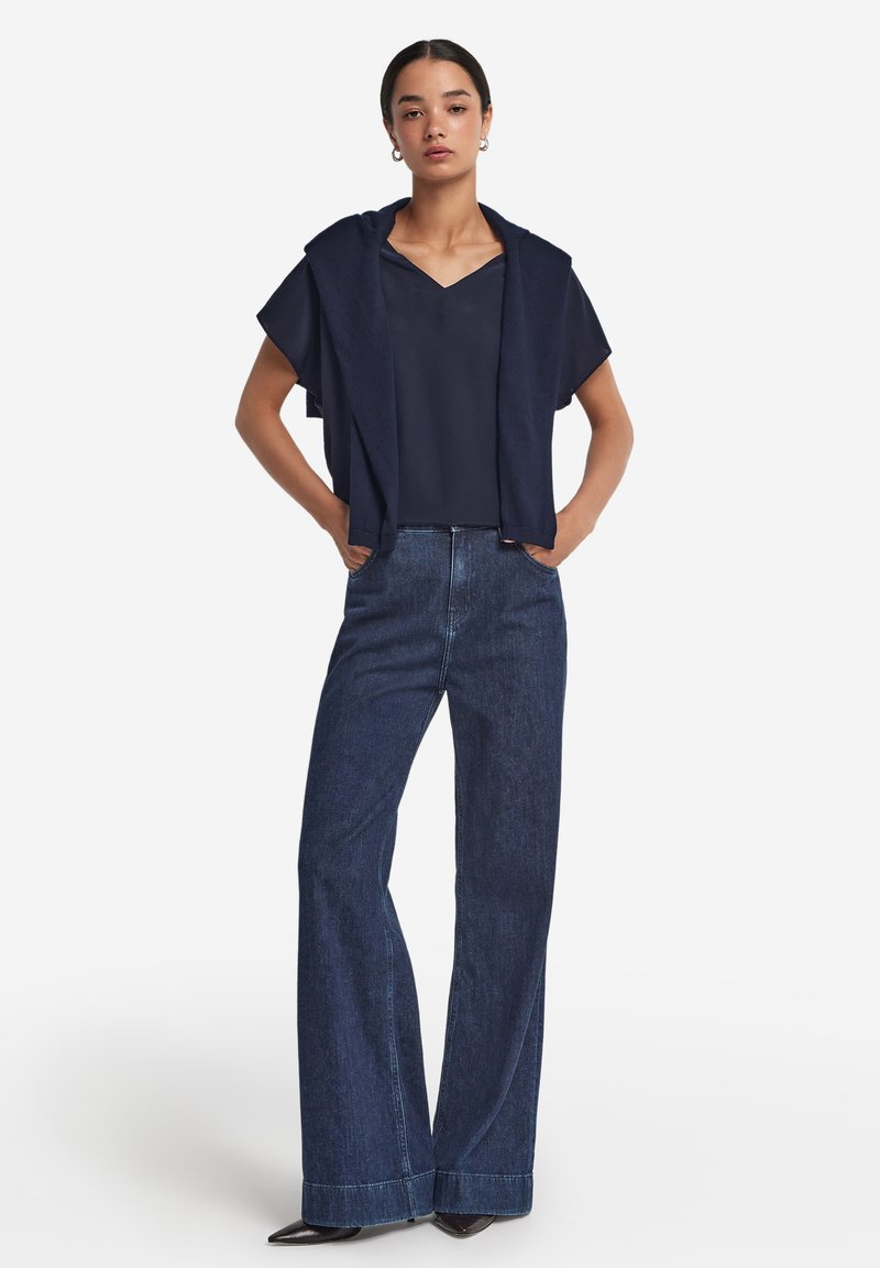 Marinblå topp med vida ärmar, lager på lager över en mörkblå skjorta, kombinerat med vida mörk denimjeans och svarta spetsiga skor.