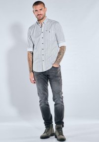 Hellgraues gemustertes Hemd mit hochgekrempelten Ärmeln, kombiniert mit dunkelgrauen Jeans und olivgrünen Stiefeln. Mit Knopfdetails und figurbetontem Design.