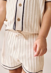 Strikket to-delt sæt i beige med teksturerede striber, korte ærmer, træknapper og navykanter. Inkluderer en sløjfedetalje ved taljen.