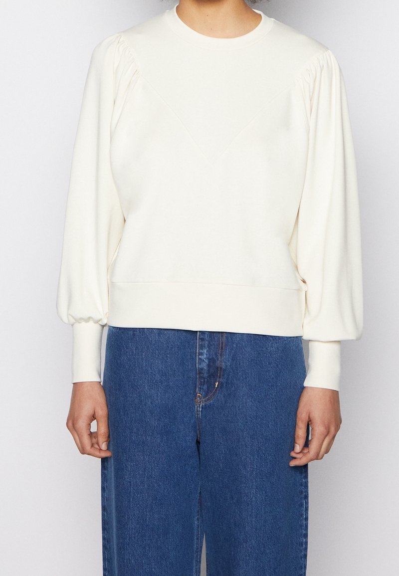 Felpa color crema con maniche a sbuffo e design a yoke a forma di V, con vita aderente e polsini a costine. Indossata con jeans blu.