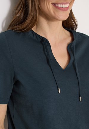 Donna sorridente con capelli castani che indossa una maglietta a V di colore verde acqua scuro con maniche corte e lacci all'altezza del collo.