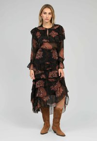 Robe noire transparente avec des manches froncées et un ourlet asymétrique, ornée d'un motif floral en paisley dans des tons terreux. Associée à des bottes de cowboy marron.