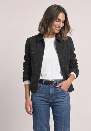 Une femme souriante porte une veste noire mouchetée sur une chemise blanche et un jean bleu, sur un fond clair et uni.