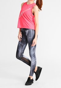 Mujer vestida con una camiseta deportiva sin mangas de color rosa brillante, leggings estampados grises de Nike y zapatillas de correr negras, de pie con una pierna ligeramente levantada.