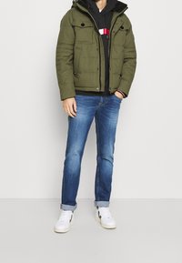 Uomo che indossa una giacca imbottita verde oliva, una felpa nera con strisce rosse e bianche, jeans blu con risvolto e sneakers bianche, in piedi contro uno sfondo bianco.