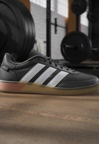 Zwart-witte Adidas sneaker met rubberen zool geplaatst op de gymvloer nabij gewichtplaten en stangen in een fitnessstudio.