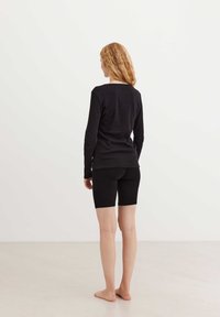 Top nero a maniche lunghe abbinato a pantaloni da ciclismo neri, con un design aderente e una texture liscia. Il modello è senza scarpe e gira di spalle.