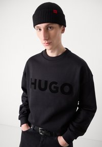 Black sweatshirt with large embossed "HUGO" logo, ribbed crew neck, paired with a black ribbed beanie featuring a red logo tag.