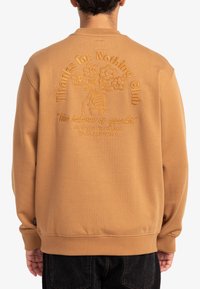 Sudadera color marrón claro con un cuello alto, que presenta texto bordado y una mano sosteniendo flores en la parte posterior. Puños y dobladillo acanalados.