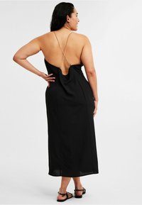 Schwarzes Maxikleid mit drapierter Rückseite, dünnen überkreuzten Trägern und einer schlanken Silhouette. Hergestellt aus glattem Stoff mit fließender Textur.