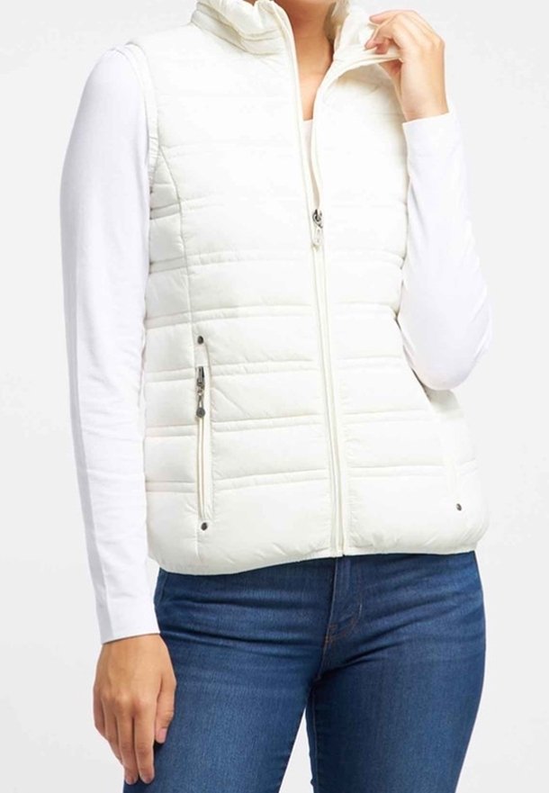 Femme portant une veste matelassée blanche sans manches avec une fermeture éclair à l'avant et des poches zippées, par-dessus un haut blanc à manches longues et un jean bleu.