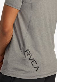 Šedé sportovní tričko s krátkými rukávy, štíhlý střih, detailní švy na bočních stranách a výrazné černé logo "RVCA" na dolním levém boku.