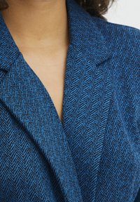 Modrý herringbone blazer s tvarovaným límcem, se strukturovanou tkaninou a hlubokým výstřihem do V. Detailní pohled na materiál.